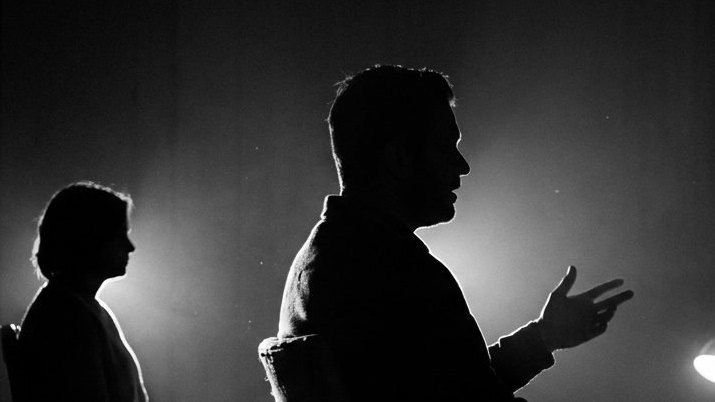 Un plateau d'interview à contre-jour : au centre, une personne de profil parle en faisant un geste ; une seconde, assise à gauche, écoute.
